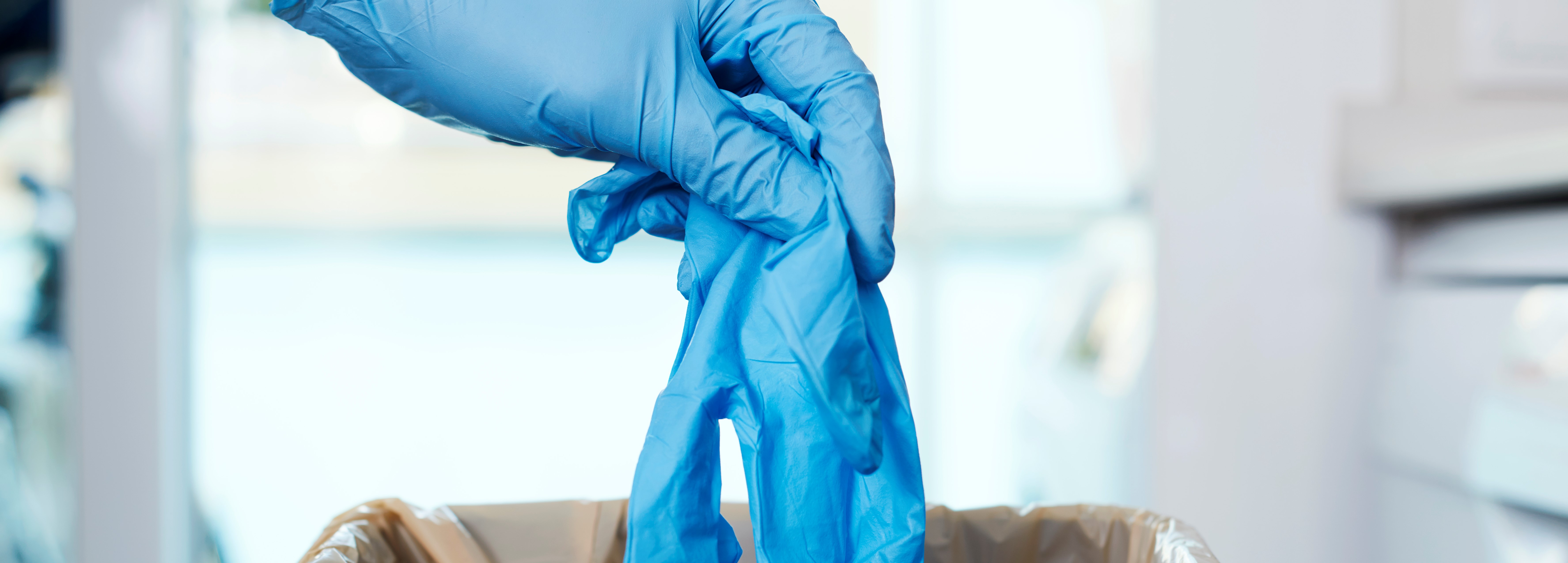 Das Bild zeigt eine Hand mit einem blauen medizinischen Handschuh an, welche einen anderen dieser Handschuhe in einen Mülleimer wirft.
