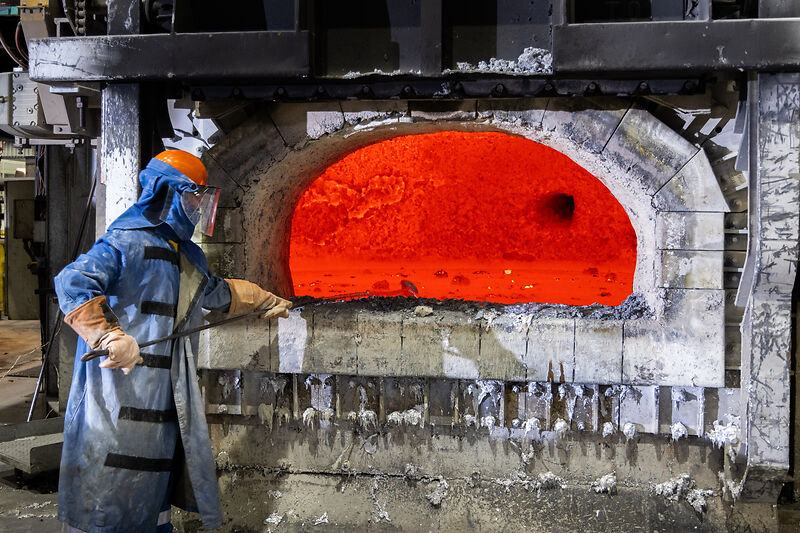 Ein Arbeiter in Schutzkleidung, einschließlich eines Helms und eines Gesichtsschutzes, steht vor einem offenen, glühend heißen Industrieofen. Der Ofen leuchtet intensiv rot und orange, was die extreme Hitze im Inneren zeigt. Der Arbeiter hält ein Werkzeug und scheint den Ofen zu überwachen. Die Umgebung ist eine industrielle Anlage.