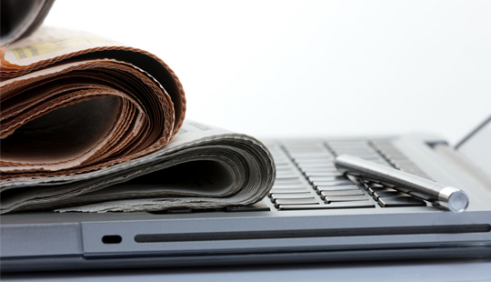 Das Foto zeigt einen aufgeklappten Laptop in Detailaufnahme. Auf der Tastatur liegen zusammengerollte Zeitungen und ein Stift.